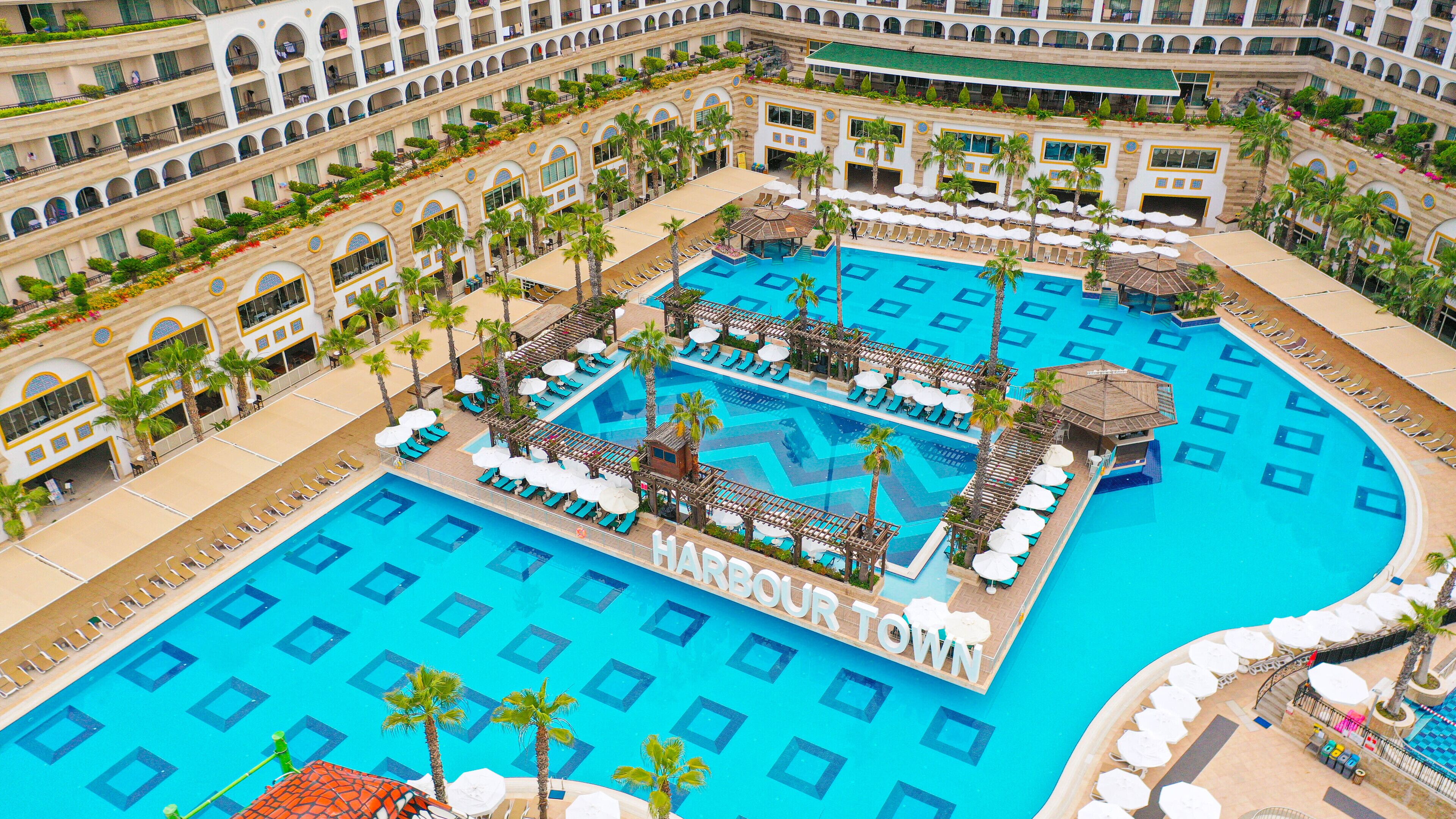 A large, multi-level hotel with a central swimming pool surrounded by sun loungers and palm trees.