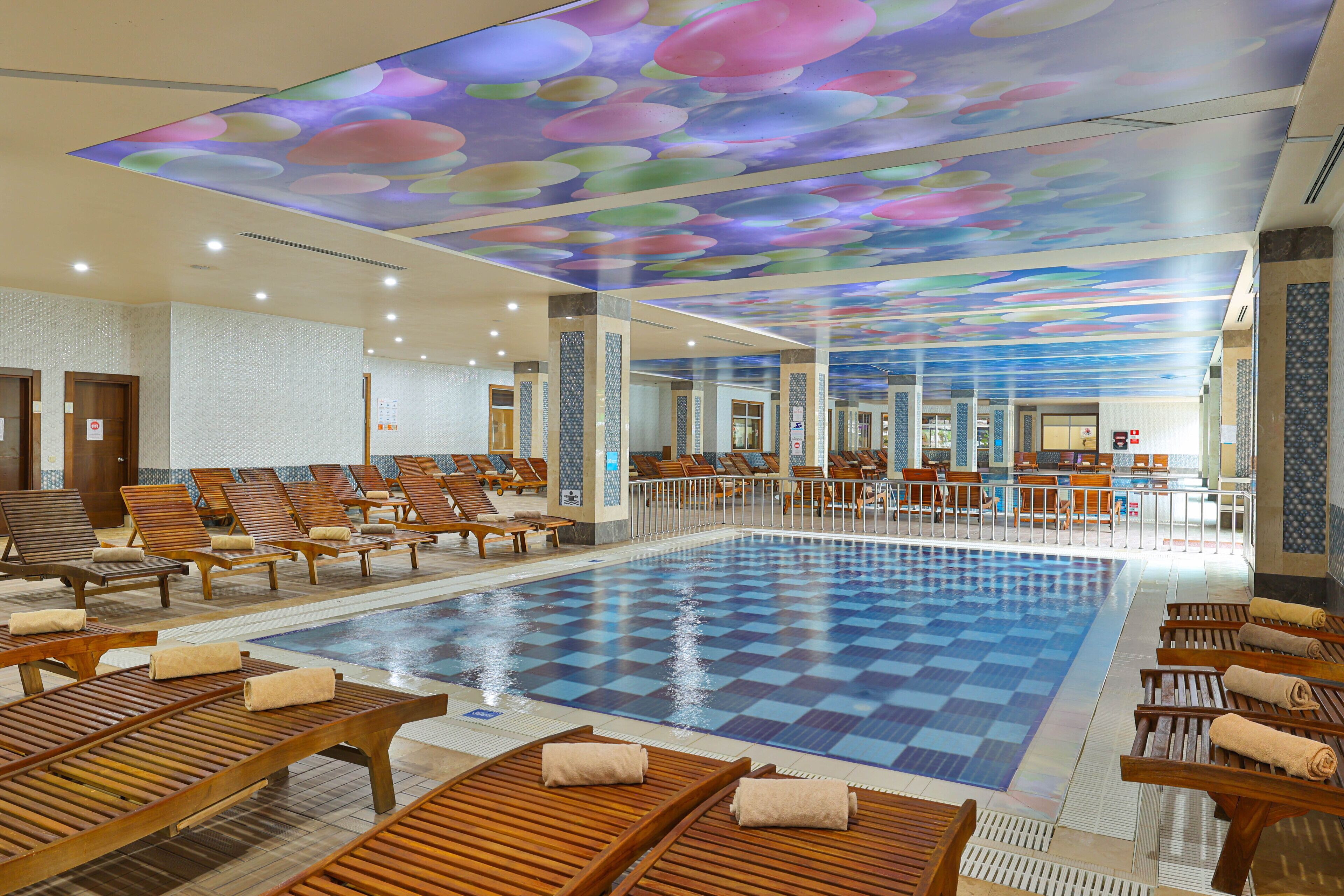 Un hall d’hôtel doté d’une grande piscine intérieure, de chaises longues et d’un plafond aux motifs colorés.