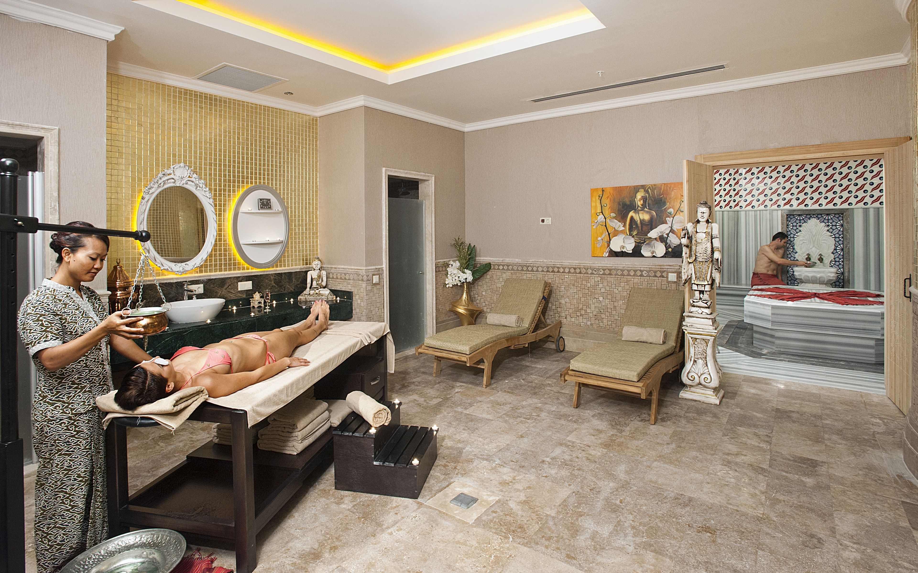 Una stanza spa con un lettino da massaggio, due poltrone relax, una statua decorativa di Buddha e un grande specchio.