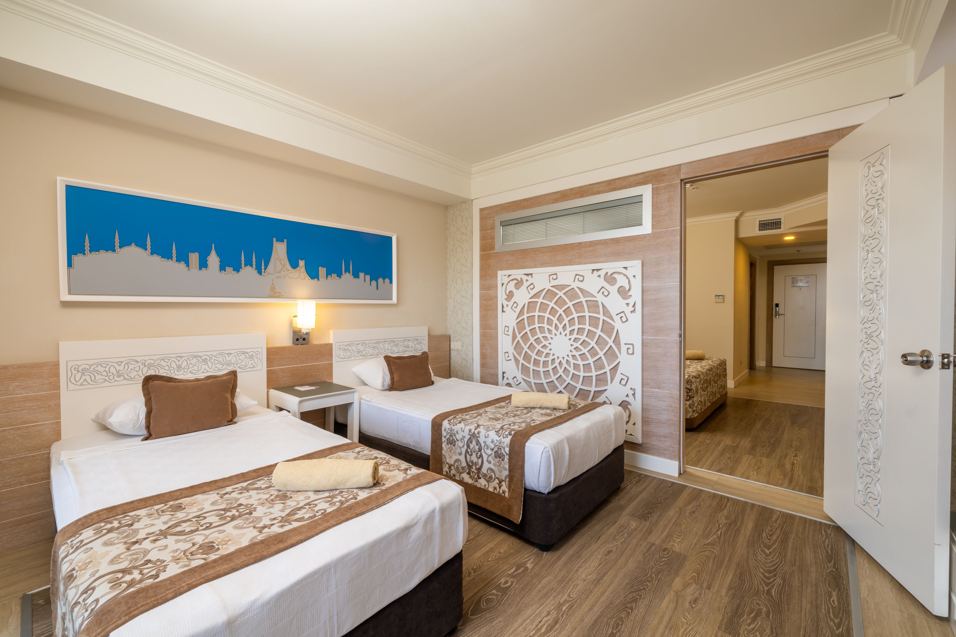 A hotel room with two beds, a cityscape painting, a decorative wall panel, and a door leading to another room.