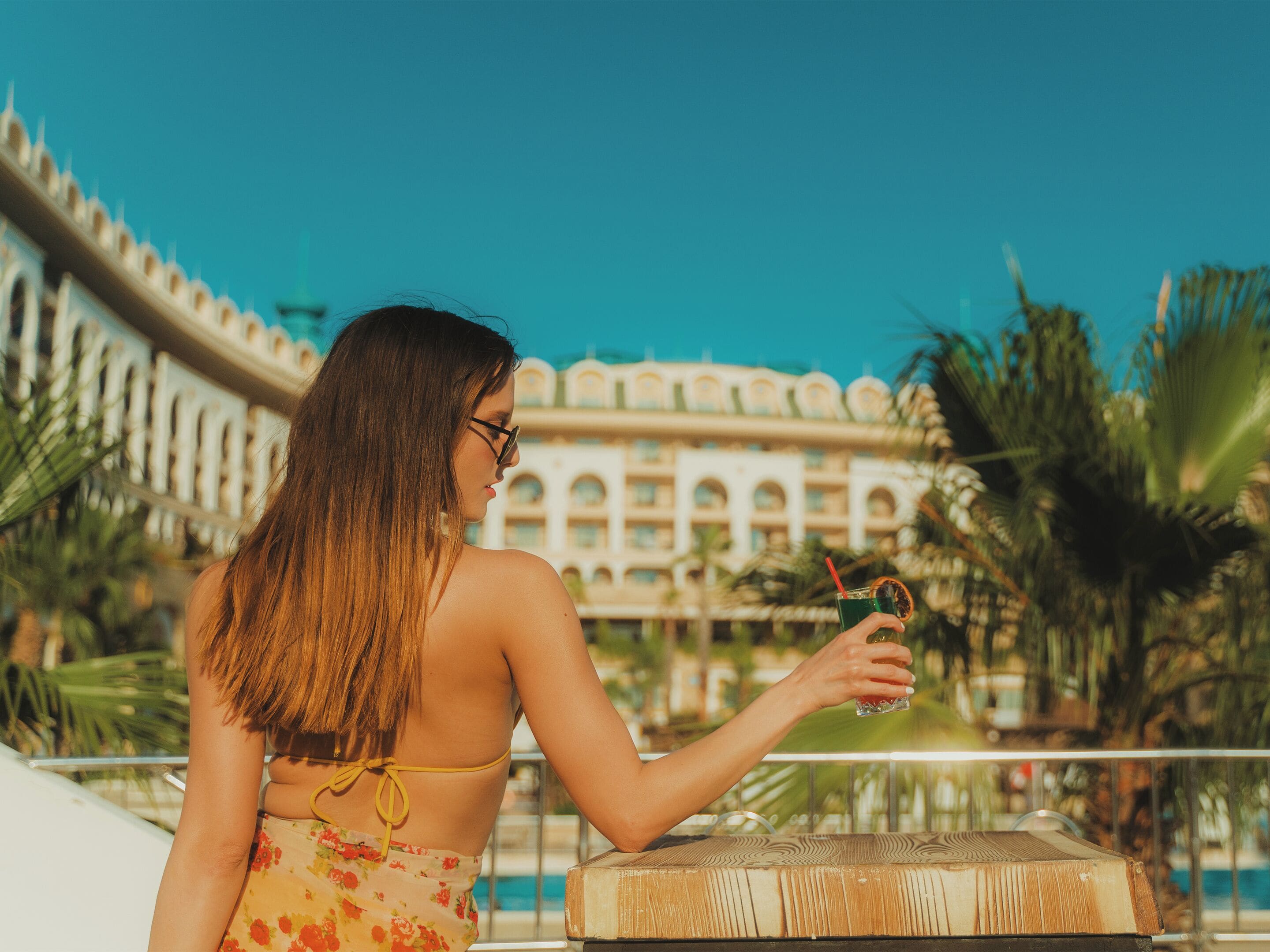 Une personne tient un verre contenant une boisson et une tranche de fruit, et se tient près d’une piscine, avec un bâtiment en arrière-plan.