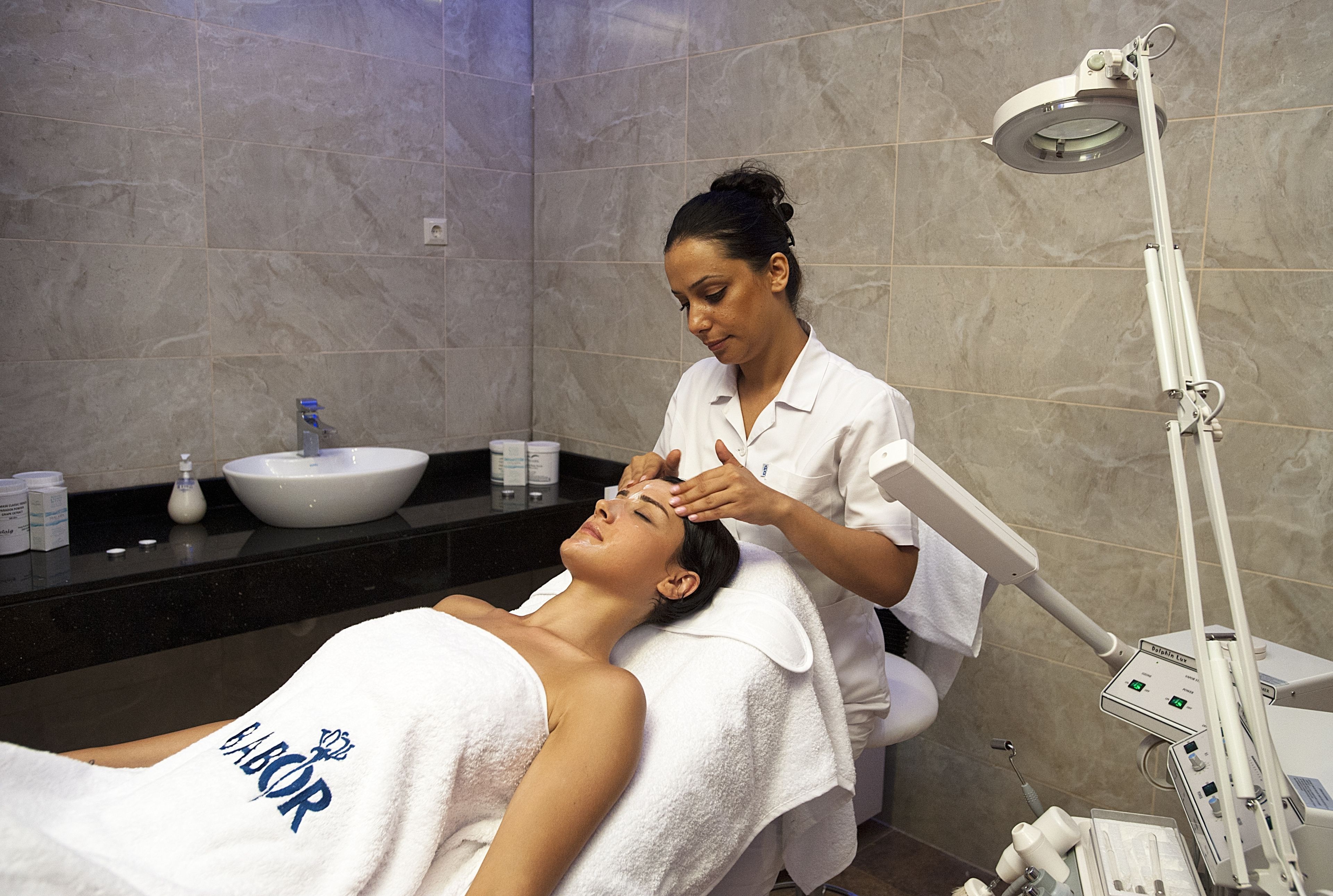A person receiving a facial treatment in a bathroom with a sink and countertop, a mirror, and a medical device.