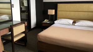 A hotel room with a bed, a desk, a chair, a nightstand with a lamp, and a mirror. - Sunny Bay Suites (Manila)