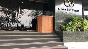 The entrance to Sunny Bay Suites, featuring a modern facade with a large logo, a wooden podium, and a planter with greenery. - Sunny Bay Suites (Manila)