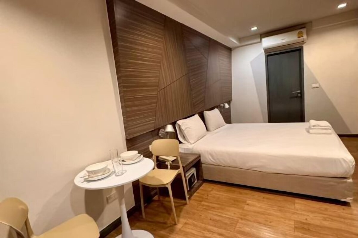 A modern hotel room with a bed, a small table with a chair, and a wall-mounted air conditioning unit.