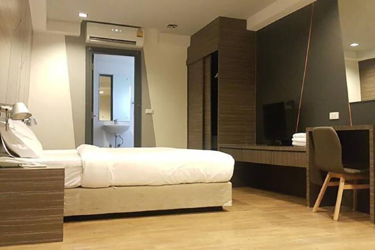 A modern hotel room with a bed, a desk with a chair, a television, and a bathroom visible through an open door.