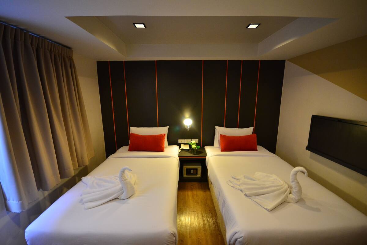 A hotel room with two beds, each with a red pillow and a white towel. There is a nightstand with a lamp between the beds.