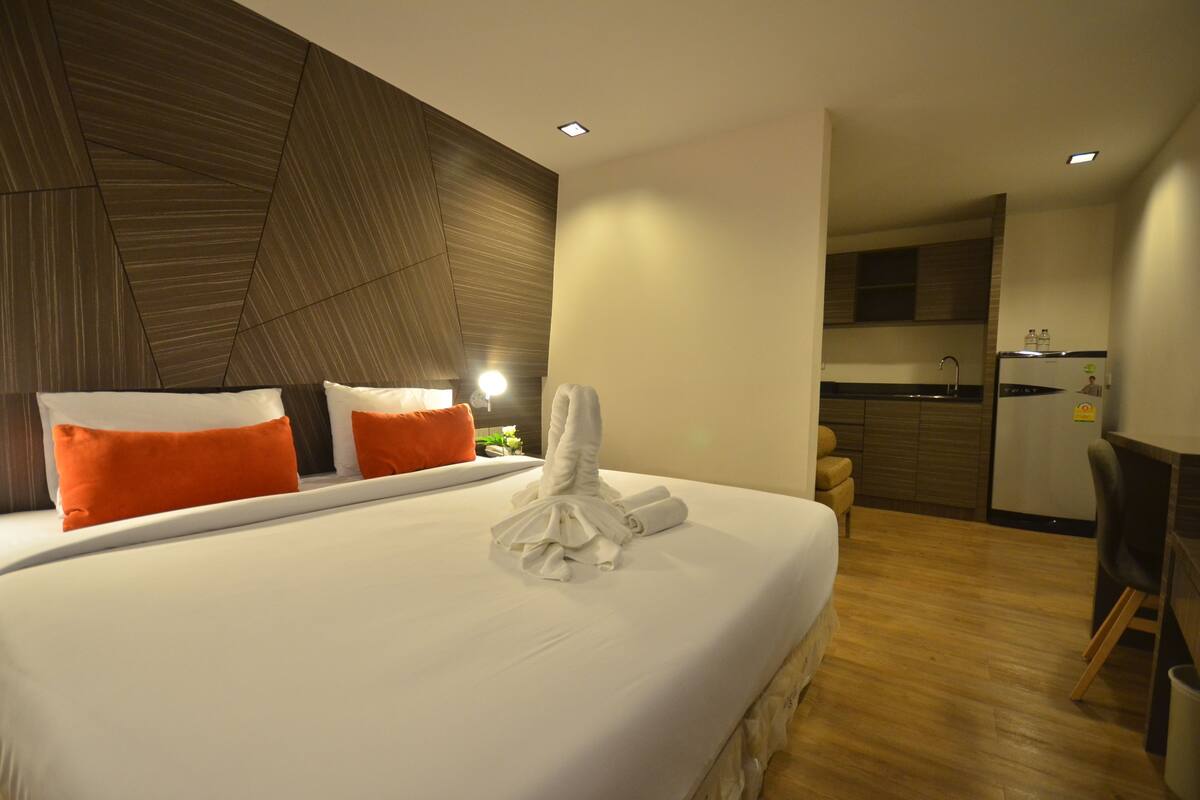 A hotel room with a large bed, orange pillows, a neatly folded white towel, a wooden accent wall, a small kitchen area, and a chair.