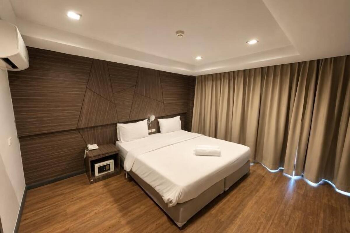 A modern hotel room with a large bed, a nightstand, a wall-mounted air conditioner, and a window with curtains.
