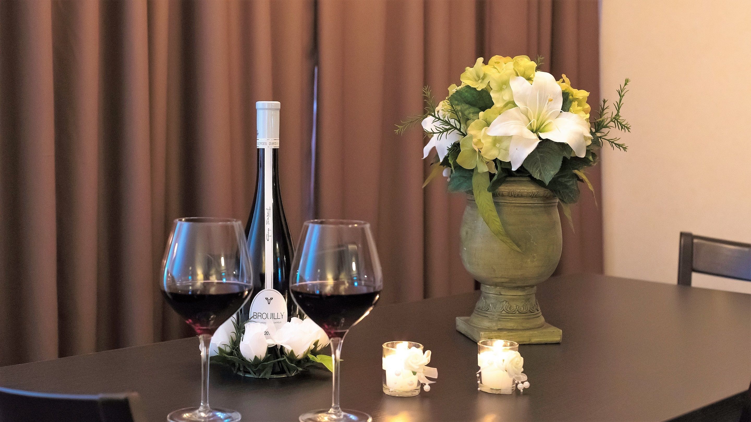 A table with two wine glasses, a bottle, a vase with flowers, and a lit candle.