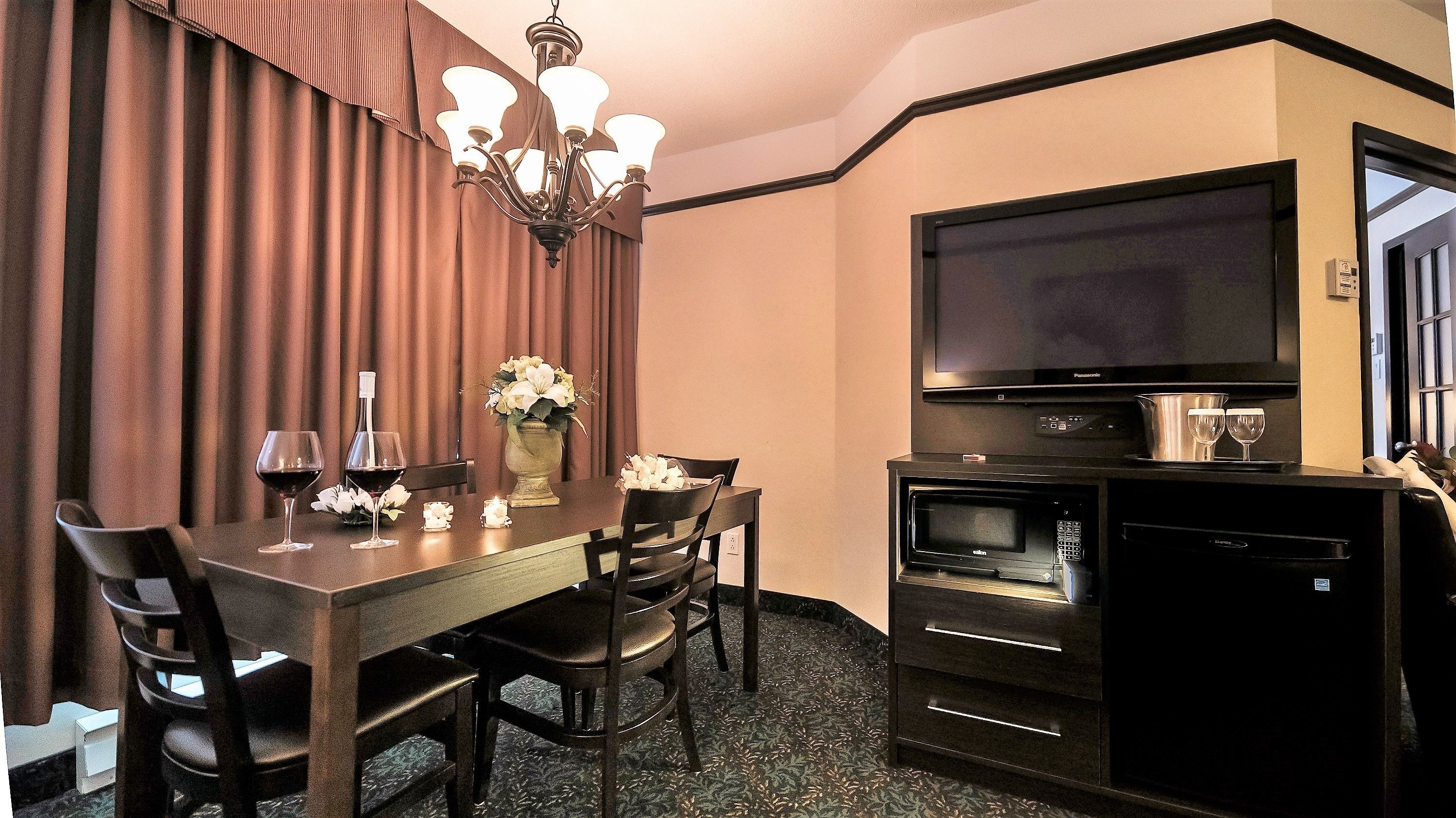 A hotel room with a dining table, chairs, a chandelier, a flat-screen TV, a microwave, and a refrigerator.