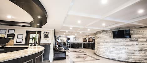 A hotel reception area with a curved counter, a staircase, and a stone wall with a television.