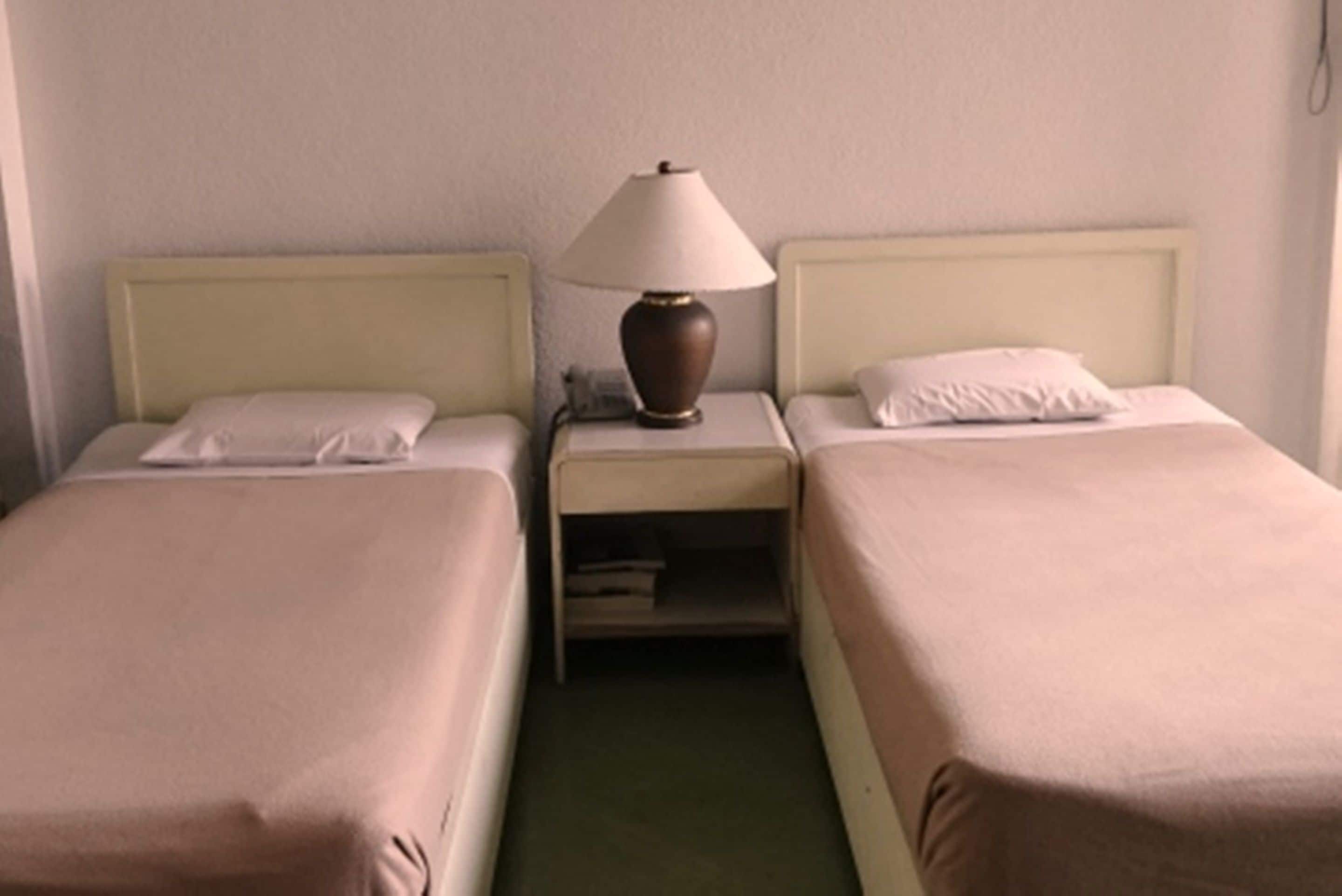A hotel room with two single beds, a nightstand with a lamp, and a small shelf.