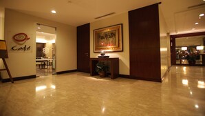 Un vestíbulo de hotel con piso de mármol pulido, una obra de arte enmarcada sobre una consola y un letrero que indica una cafetería.