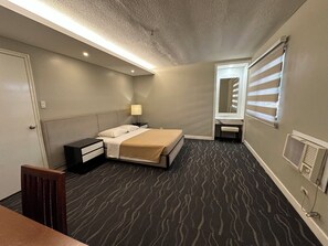 A hotel room with a bed, bedside table, lamp, and a window with blinds. - Amorsolo Mansion (Makati)