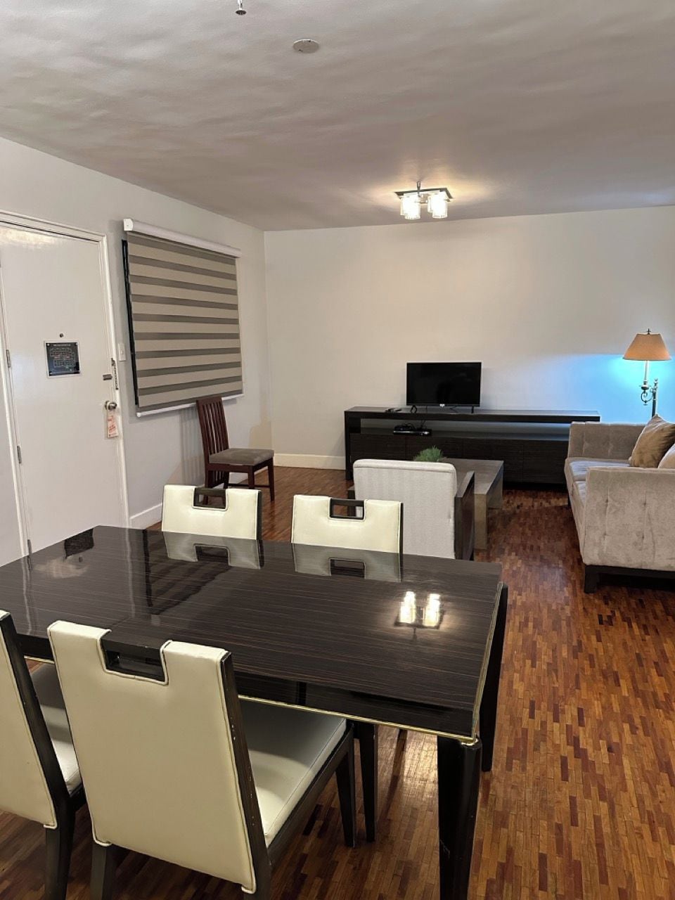 A modern living room with a dark wooden dining table, white chairs, a sofa, and a TV.