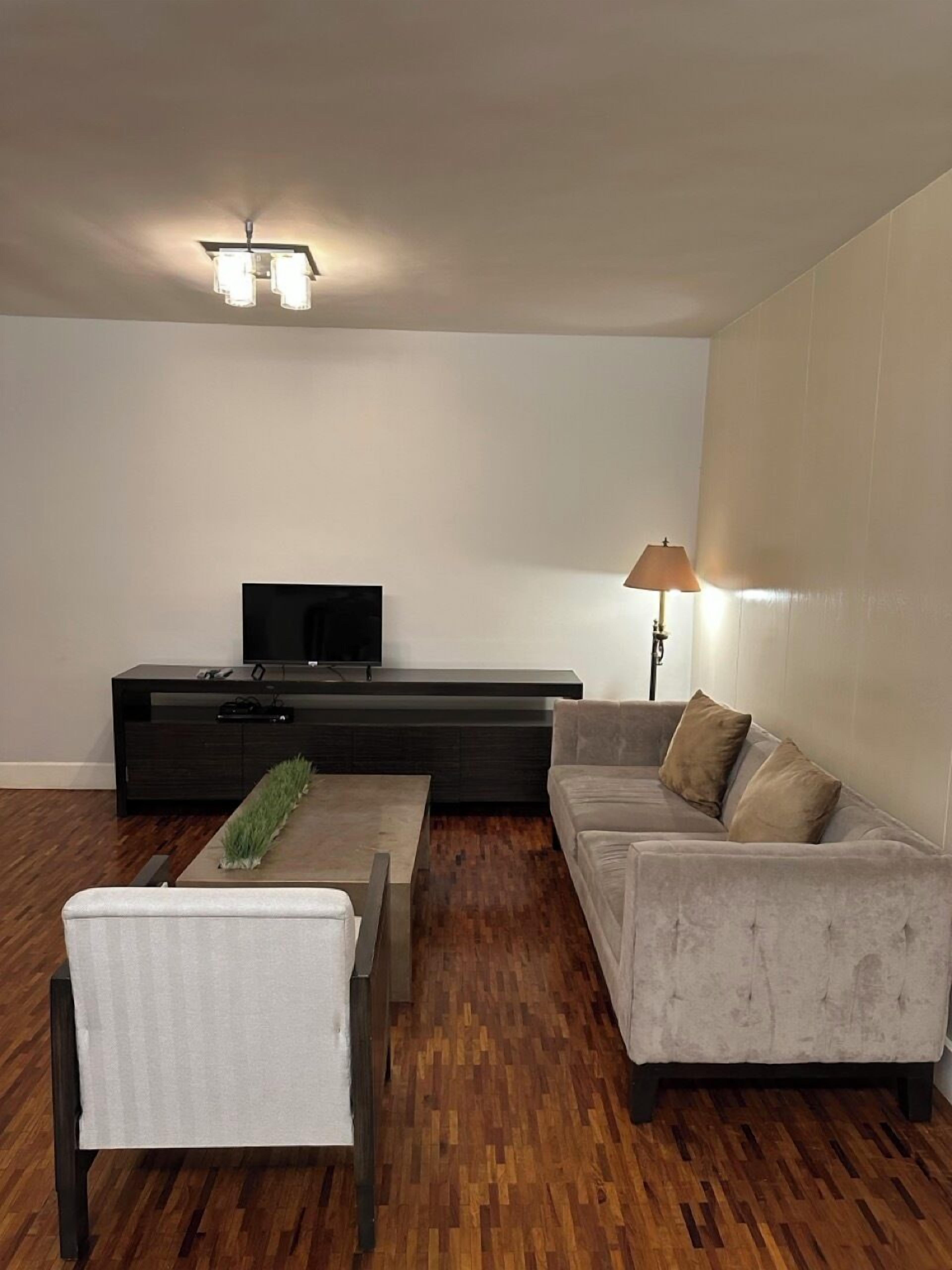 A modern living room with a flat-screen TV on a black entertainment unit, a grey sofa, a white armchair, and a wooden coffee table.