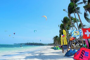 A beach with clear skies, people kite surfing, and colorful flags.
