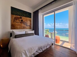 Un dormitorio con una cama grande, un cuadro en la pared y vista al océano a través de un ventanal corredero.