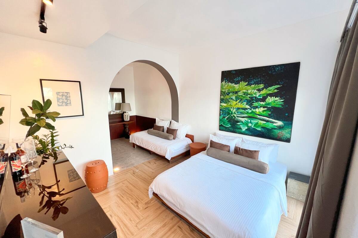 Une chambre d’hôtel avec un grand lit, un lit plus petit, un bureau avec une plante et une grande fenêtre.