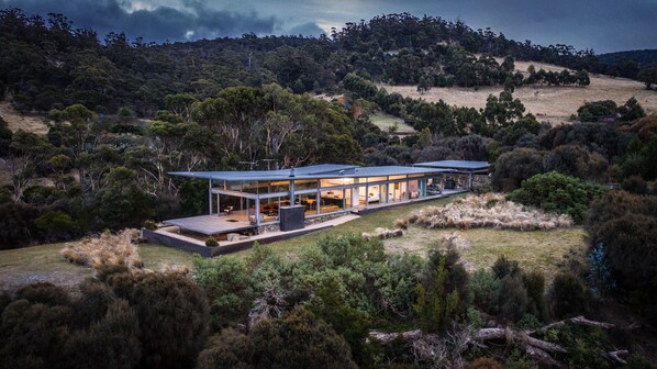 A modern house with a glass facade, large windows, and a flat roof, surrounded by dense vegetation and hills.