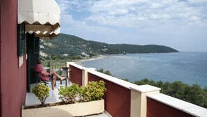Varanda com vista para uma praia e o mar, uma pessoa sentada numa cadeira e um vaso com planta.