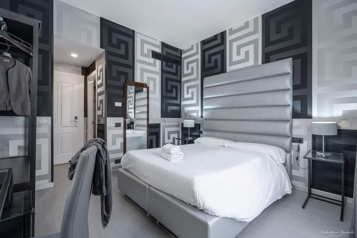 Una habitación de hotel moderna con una cama grande, un armario, un espejo y una mesita de noche con lámpara.