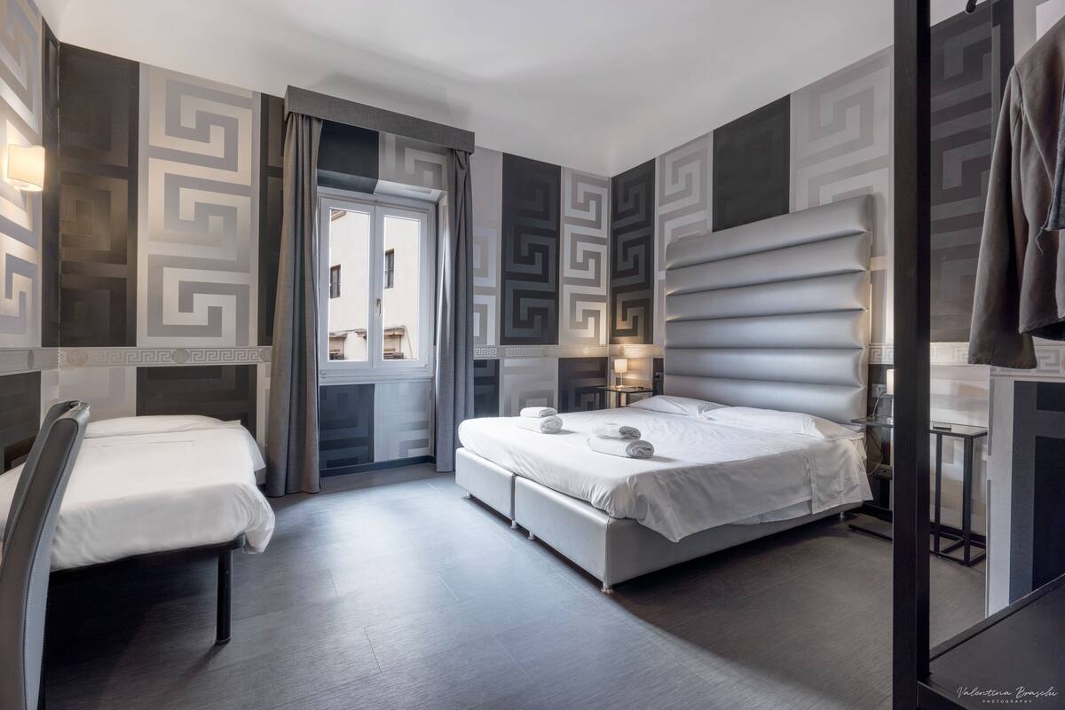 Una habitación de hotel moderna con una cama grande, una mesita de noche, una silla y un ventanal con cortinas.