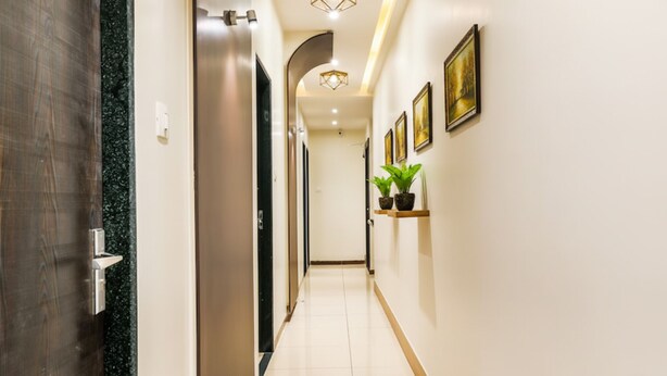 Un couloir avec une porte en bois, un mur noir et un sol carrelé de carreaux blancs. On y trouve deux plantes en pot sur une étagère et plusieurs tableaux encadrés accrochés au mur.