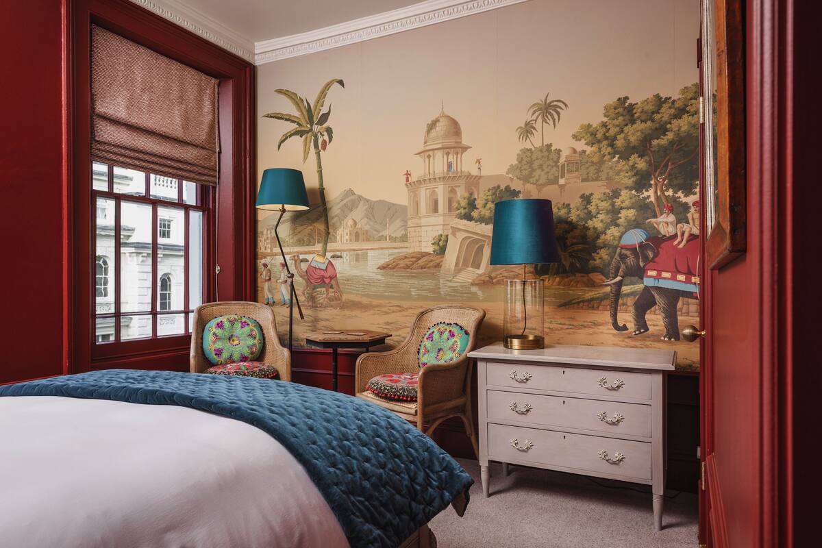 A hotel room with a bed, two chairs, a dresser, and a mural of a historical building.