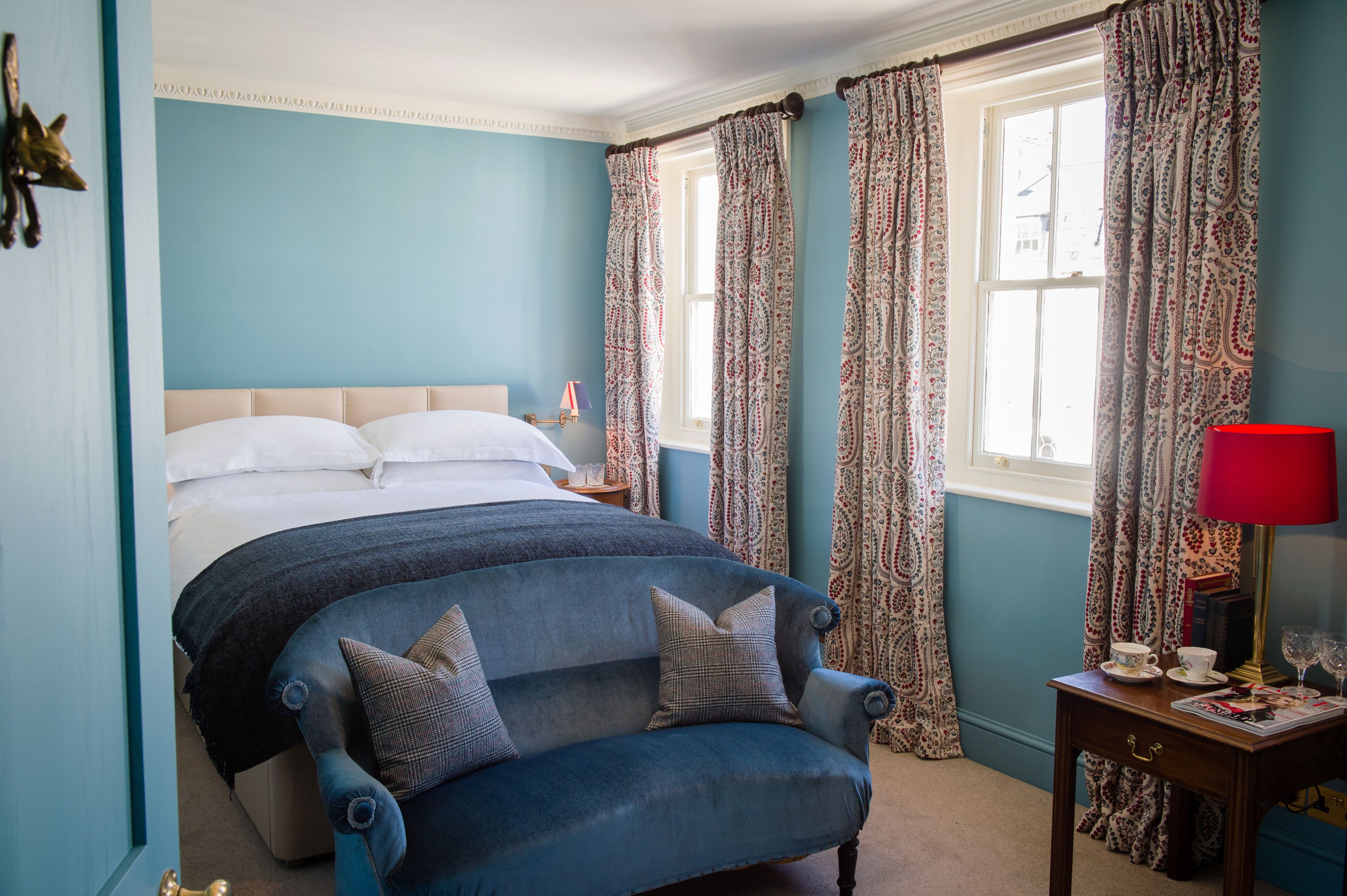 A bedroom with a bed, a blue armchair, a nightstand with a lamp, and patterned curtains.