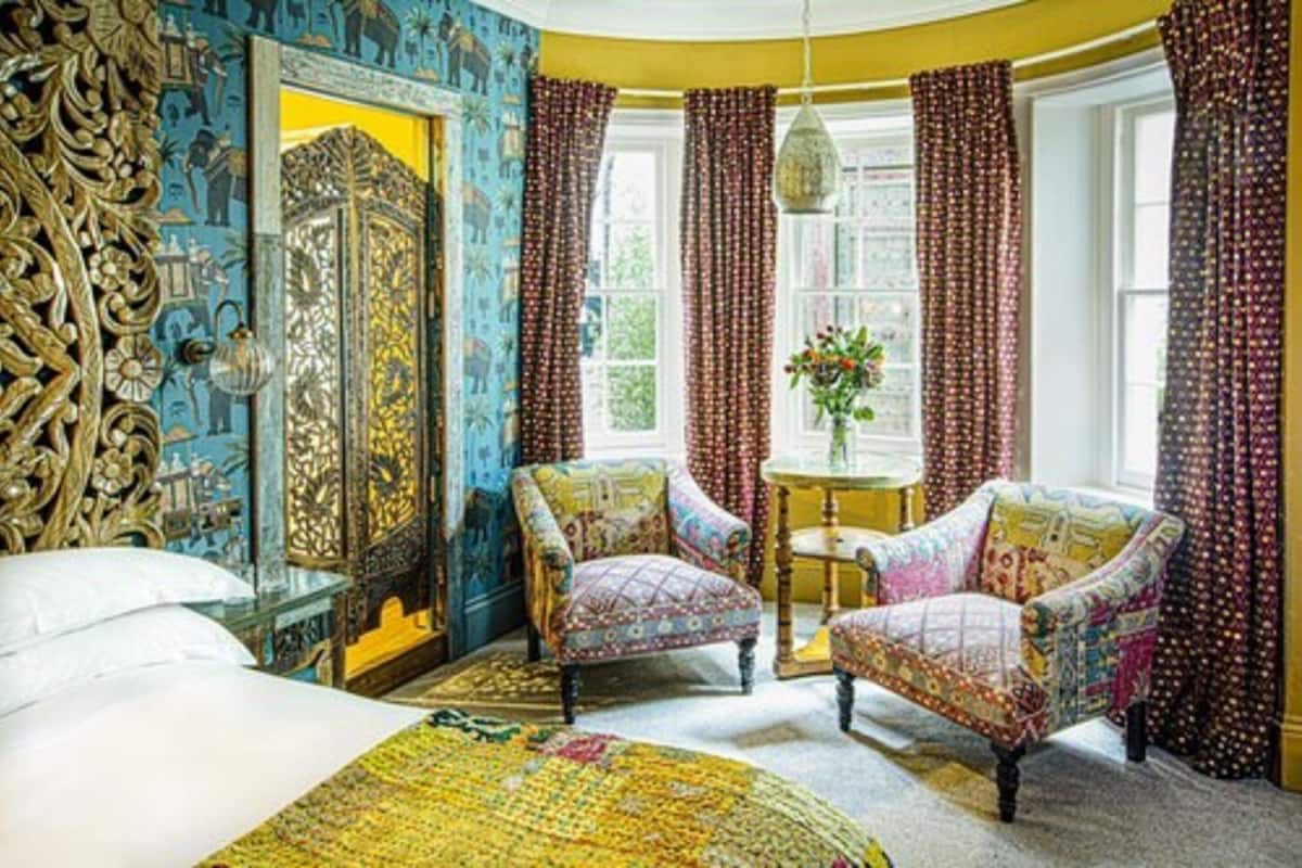 A room with a bed, two patterned armchairs, a small round table with a vase of flowers, and a large ornate mirror.