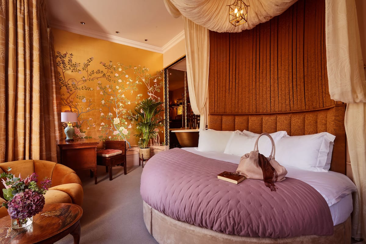 A hotel room with a large bed, a sitting area with a chair and a table, a nightstand with a lamp, and a wall with floral wallpaper.