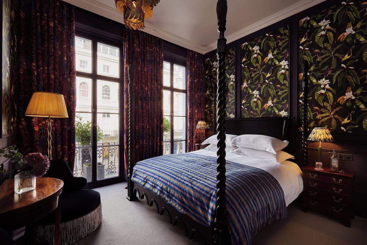 A four-poster bed with a floral-patterned headboard, a round wooden table with a vase of flowers, and a view of a building through large windows with patterned curtains.