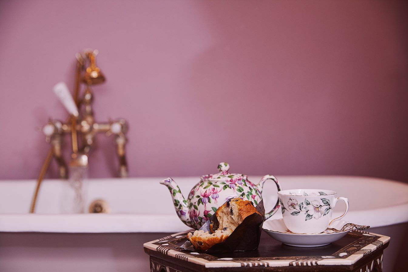 Une salle de bain avec une baignoire, une théière à motifs floraux, une tasse et un muffin sur un plateau.