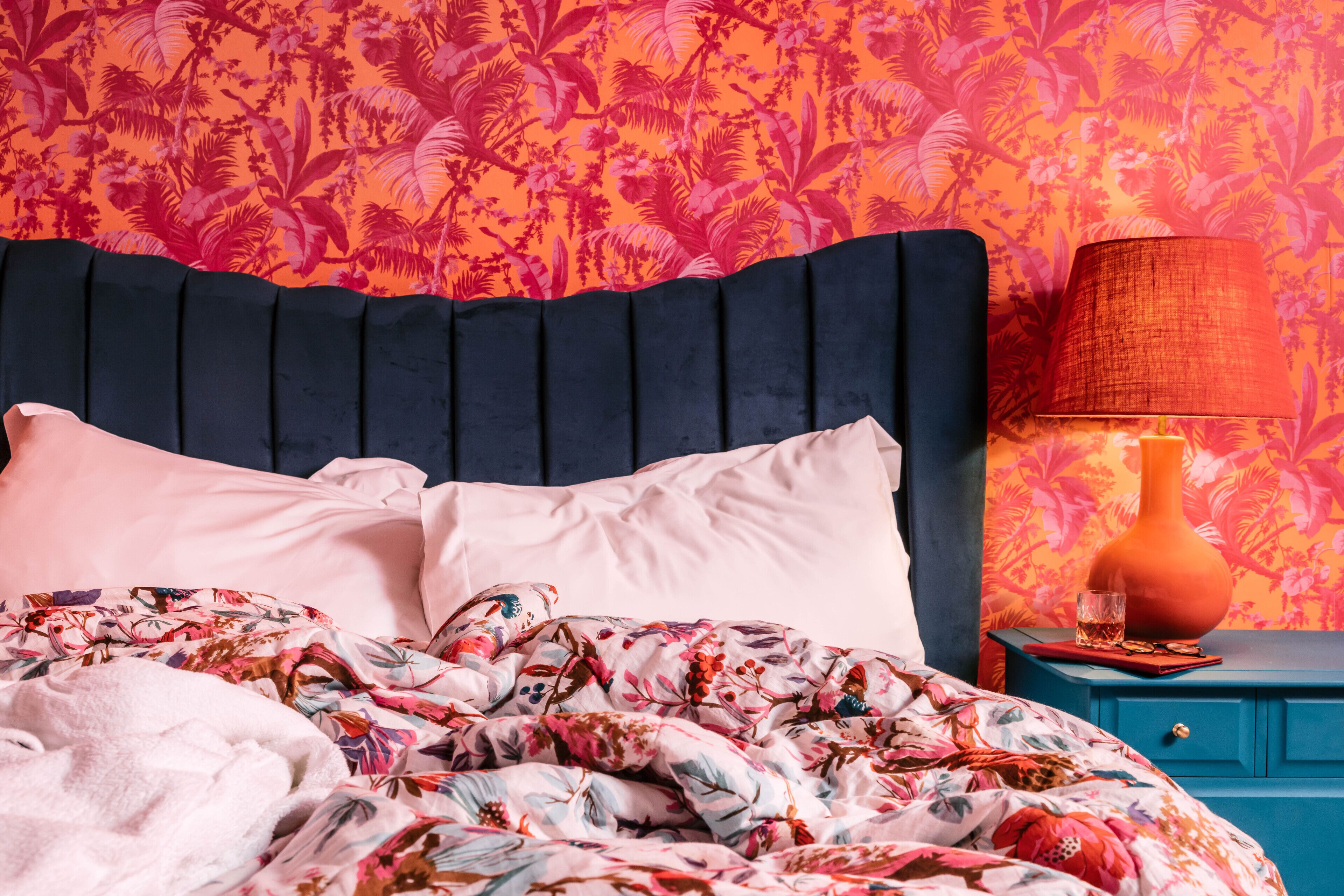 Ein Bett mit einer blumigen Tagesdecke, einem dunklen Kopfteil und einem Nachttisch mit Lampe und einem Glas Wasser.