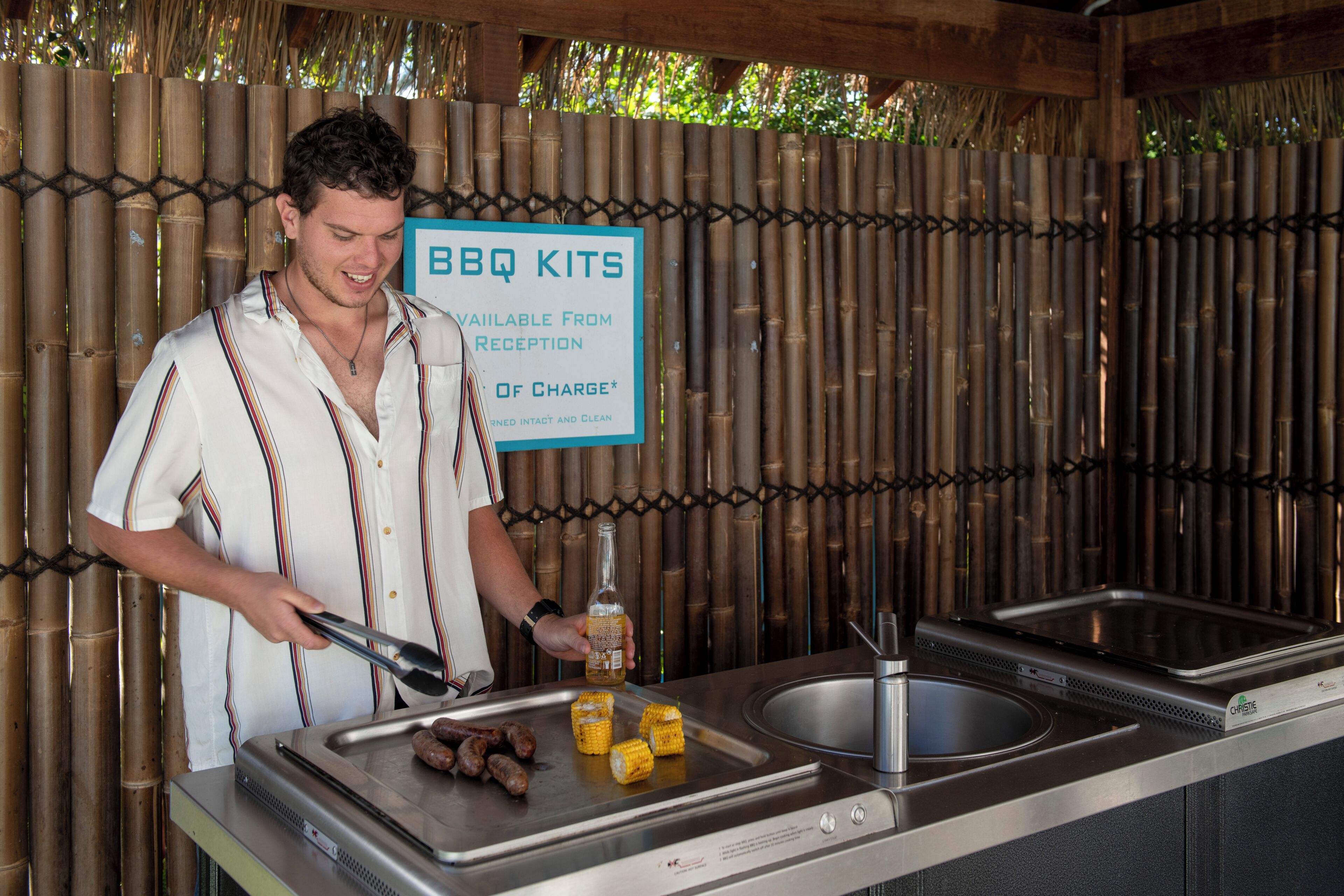 A person grilling sausages and corn on a BBQ grill with a bamboo backdrop and a sign indicating BBQ kits are available.