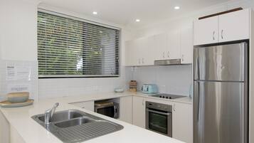 A modern kitchen with white cabinets, stainless steel appliances, a built-in oven, and a window with blinds.
