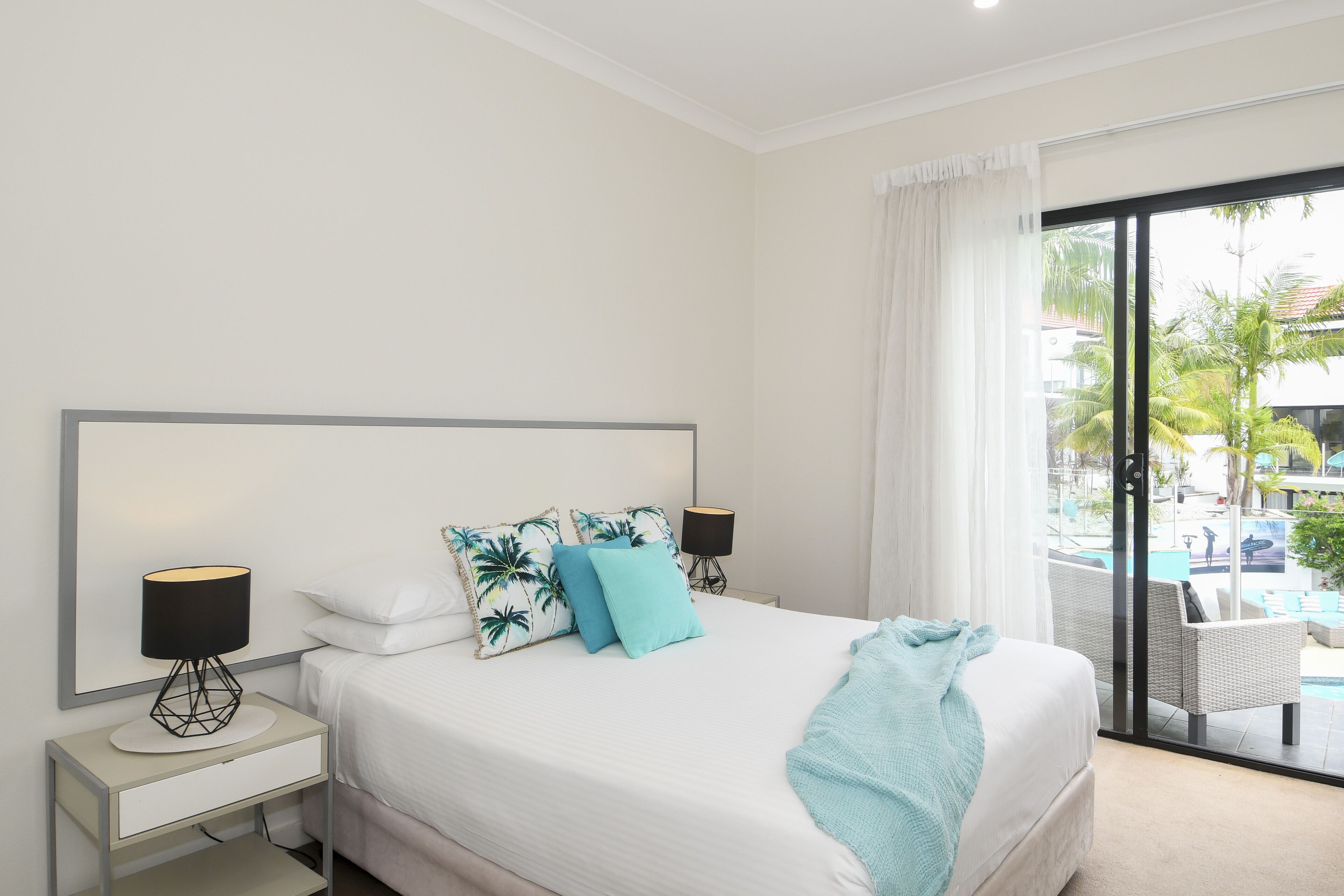 A hotel room with a large bed, two bedside tables, a nightstand, a window with a view of a pool and palm trees, and a balcony with outdoor furniture.