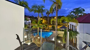 Zona de piscina con barandillas de vidrio, palmeras y un edificio de techo rojo al fondo.