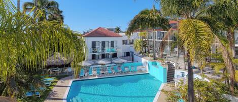 A pool area with lounge chairs and umbrellas, surrounded by palm trees and a building with a red roof.
