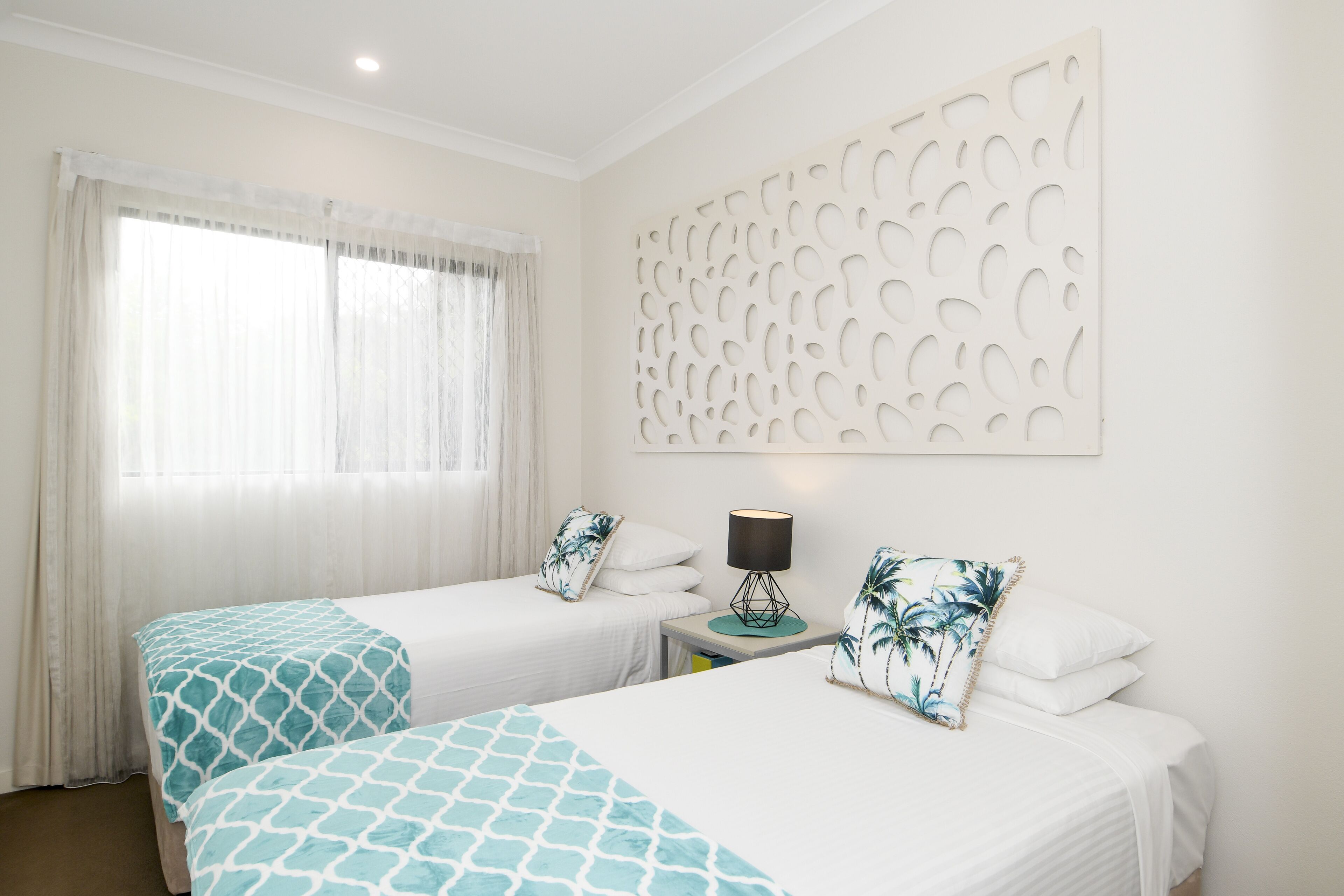 A hotel room with two beds, a window with sheer curtains, a wall-mounted decorative panel, and a small table with a lamp.