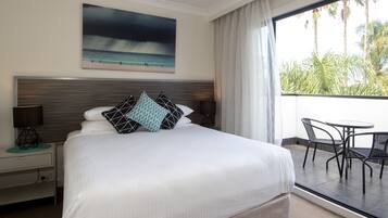 Habitación de hotel con una cama grande, un cuadro de una playa, un balcón con mesa y sillas, y vistas a palmeras.