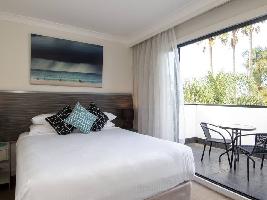 Habitación de hotel con una cama grande, un cuadro de una playa, un balcón con mesa y sillas, y vistas a palmeras.