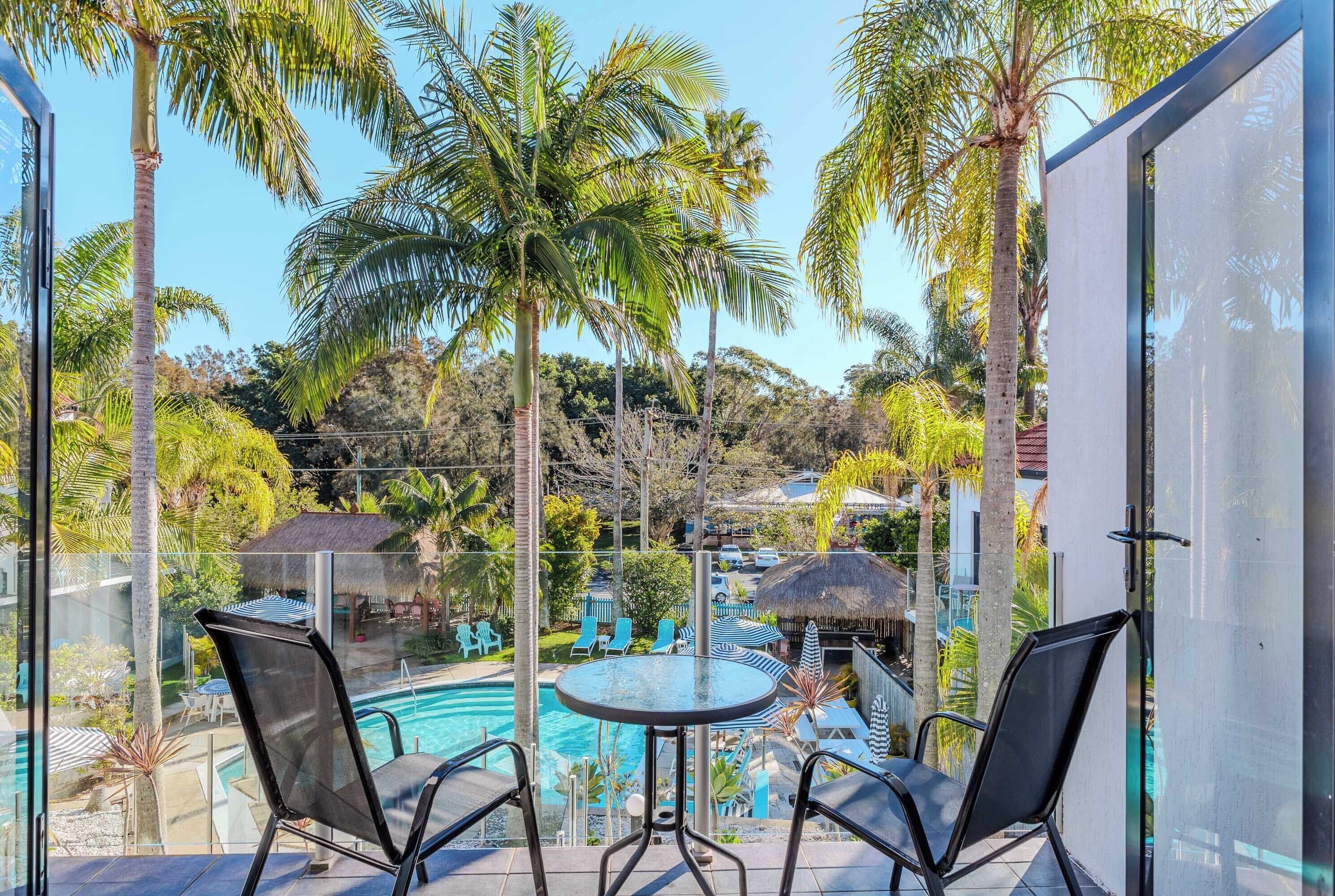 Un balcon avec deux chaises et une table donnant sur un espace piscine agrémenté de chaises longues et de parasols, le tout entouré de palmiers et offrant une vue sur un complexe hôtelier.