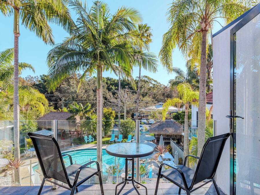 Een balkon met twee stoelen en een tafel, met uitzicht op een zwembadgedeelte met ligbedden en parasols, omgeven door palmbomen en een uitzicht op een resort.