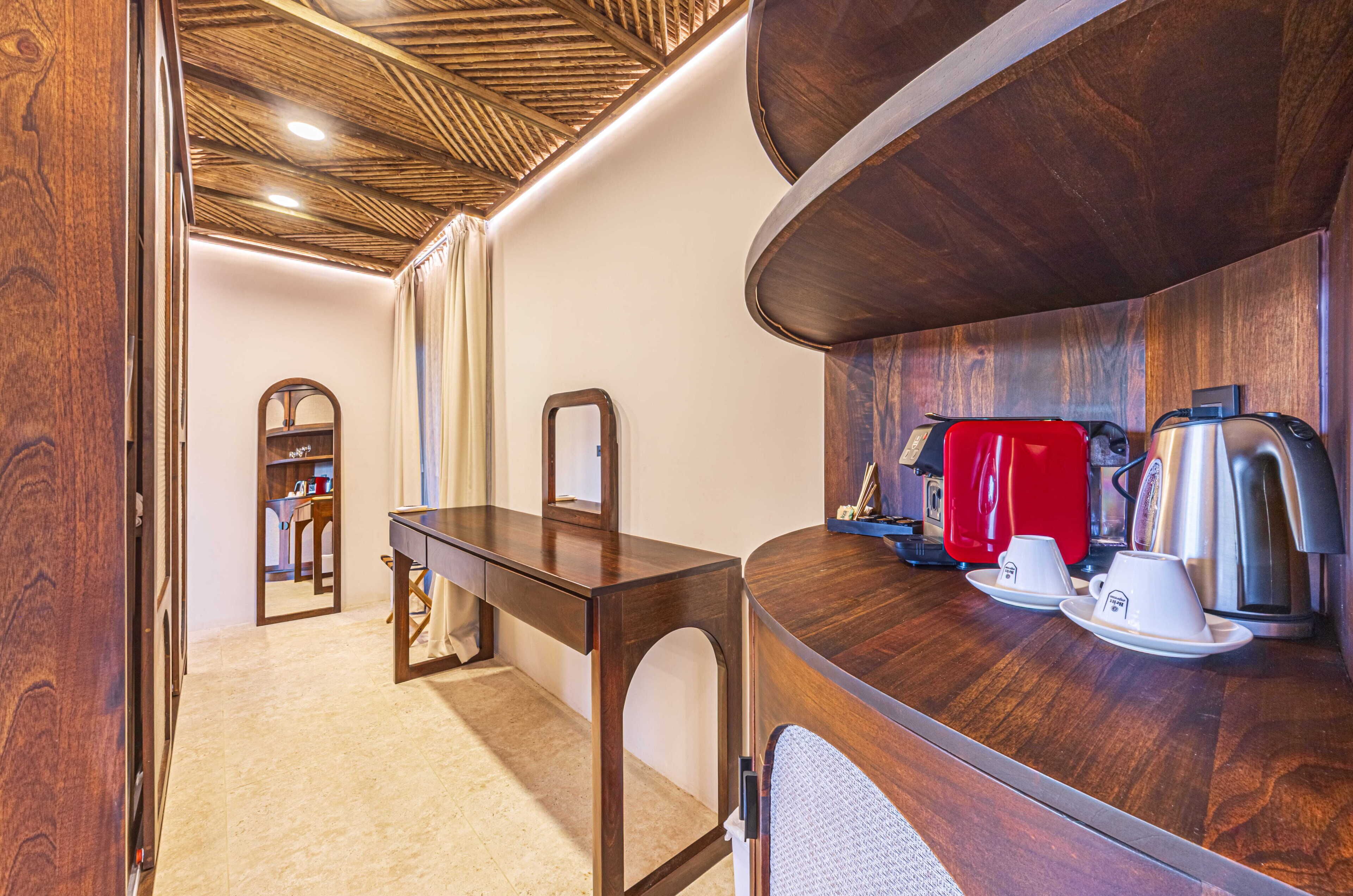 A compact room with a wooden ceiling, a desk, a mirror, a red coffee maker, and a kettle.