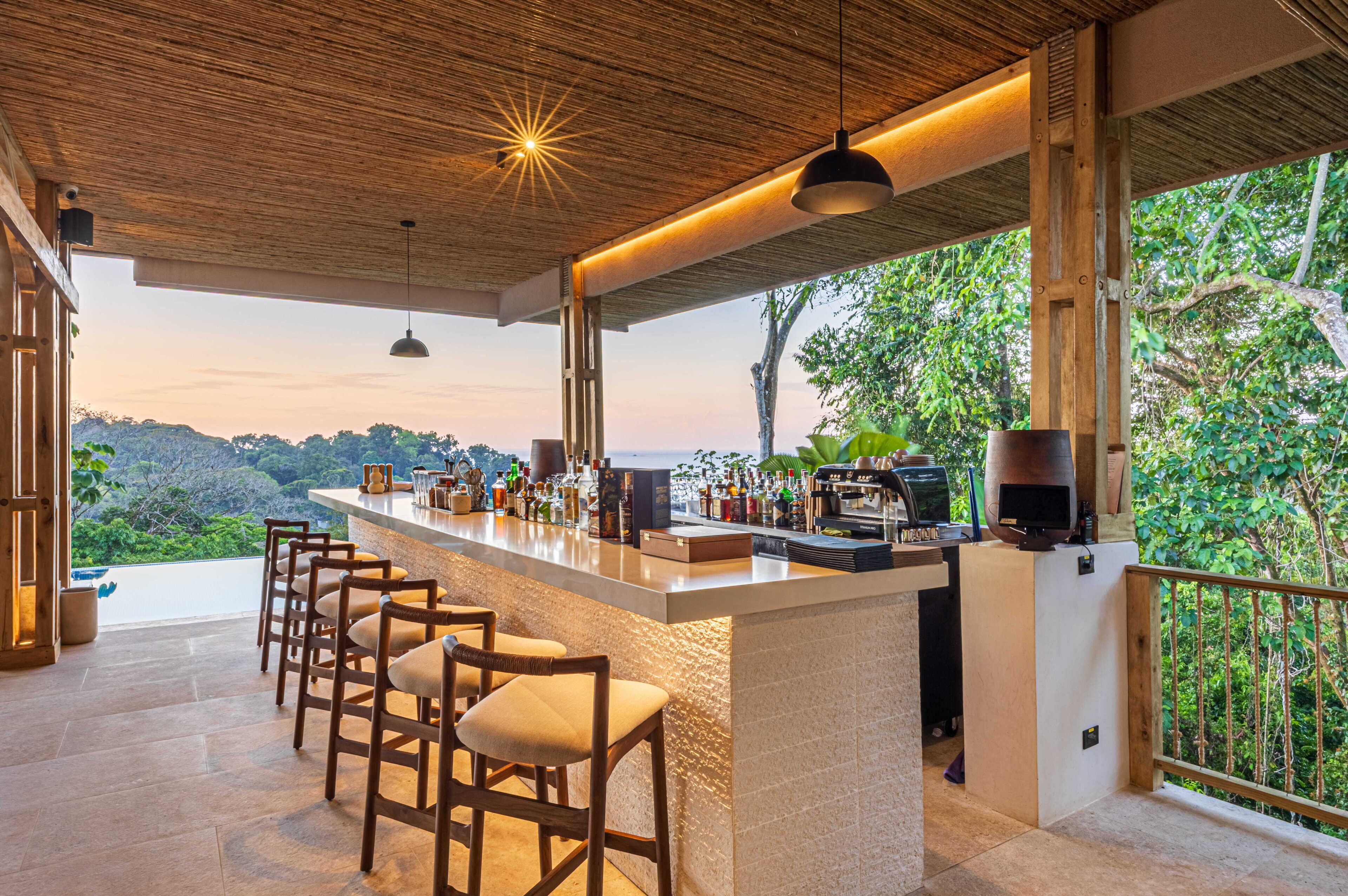 A bar area with a wooden ceiling, bar stools, and a view of the forest.