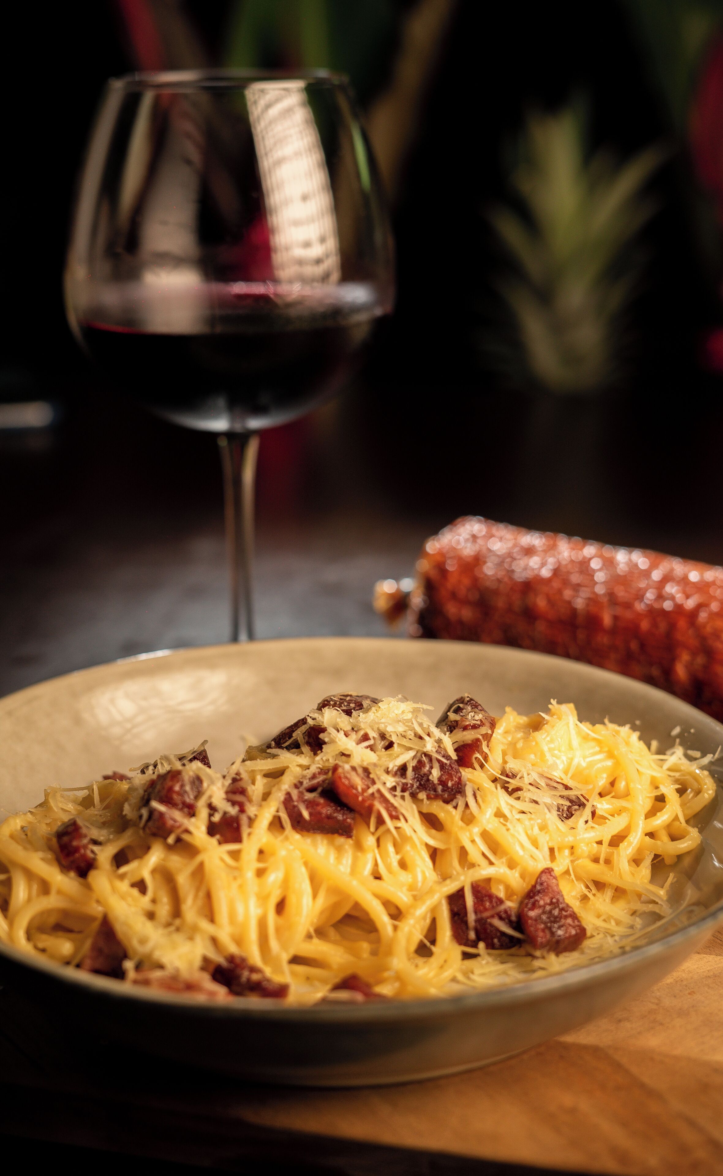 A plate of spaghetti with cheese and meat, a glass of red wine, and a sausage on a wooden table.