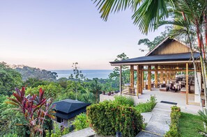 Un resort con una zona de estar al aire libre techada, abundante vegetación y vista al océano.